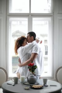 loving ethnic couple hugging and kissing while standing near window at home